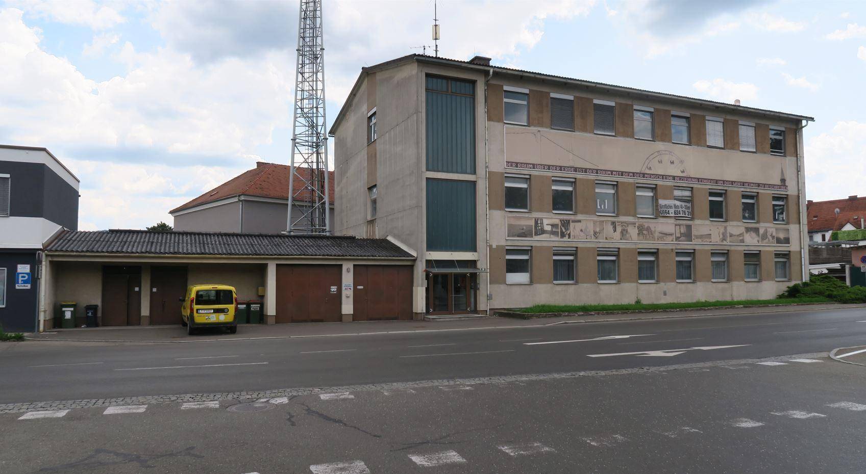 Gebäude mit beiger Fassade und angrenzenden Garagen, ein Mobilfunkmast ragt in den bewölkten Himmel.