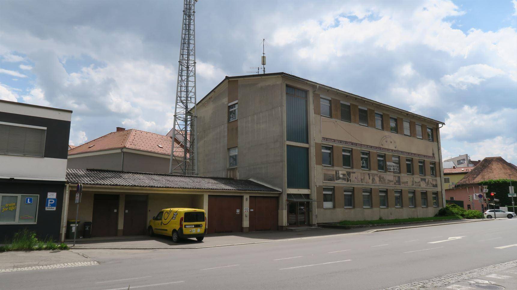 Ansicht eines mehrstöckigen Gebäudes mit angrenzender Garage und einem Mobilfunkmast im städtischen Umfeld.