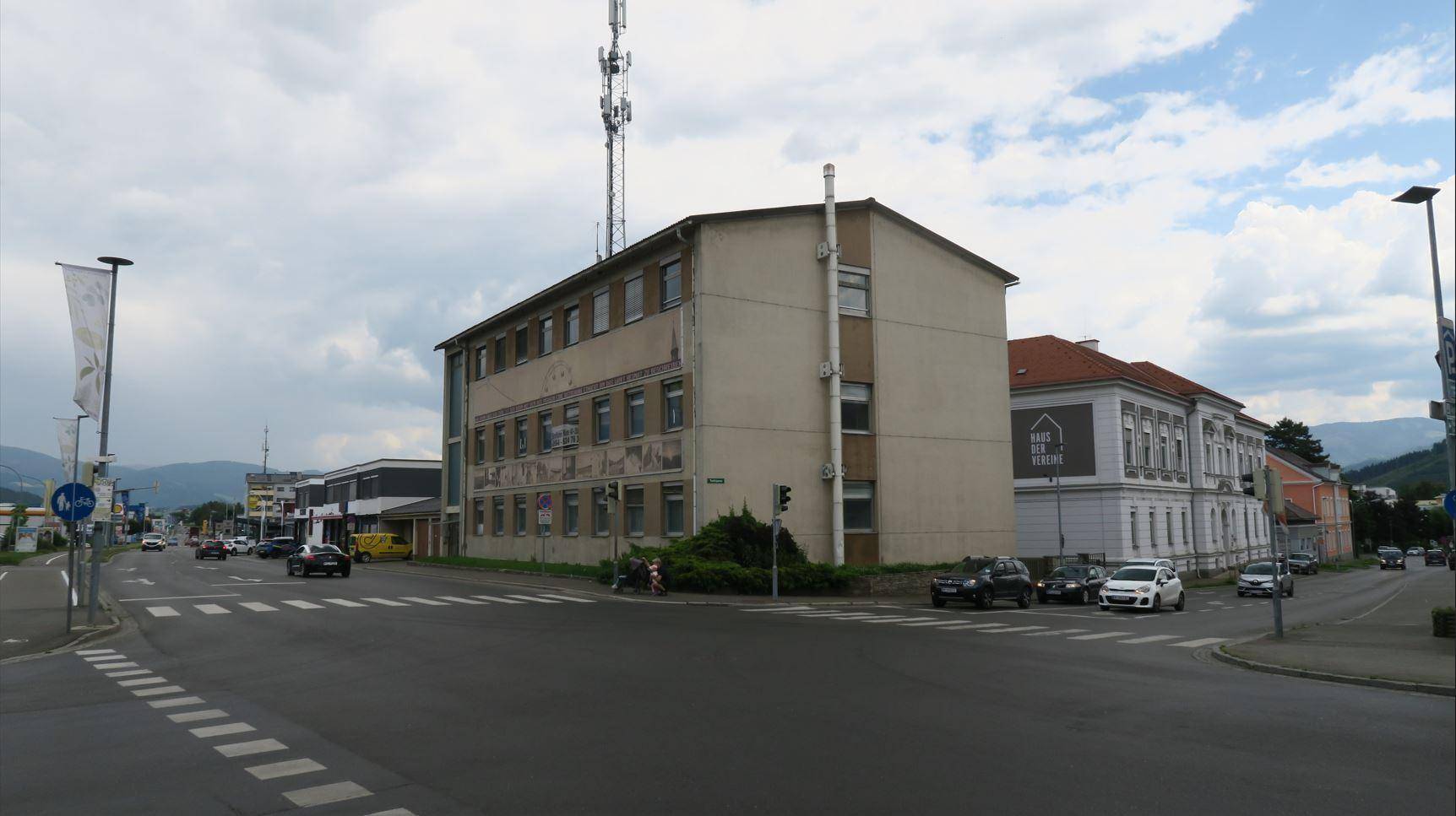 Eckgebäude an einer belebten Kreuzung mit Mobilfunkmast und umliegender städtischer Bebauung unter bewölktem Himmel.