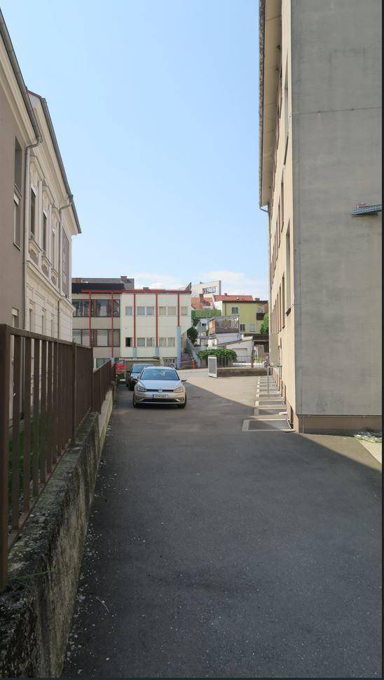 Schmale Gasse zwischen Gebäuden mit geparkten Autos und Blick auf weitere Häuser in der Ferne.