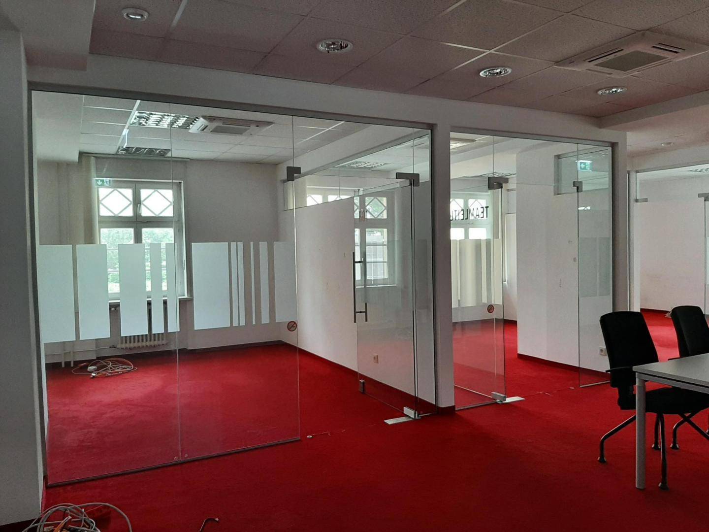 Großer Büroraum mit rotem Teppichboden, Glaswänden und einem Konferenztisch mit Stühlen.
