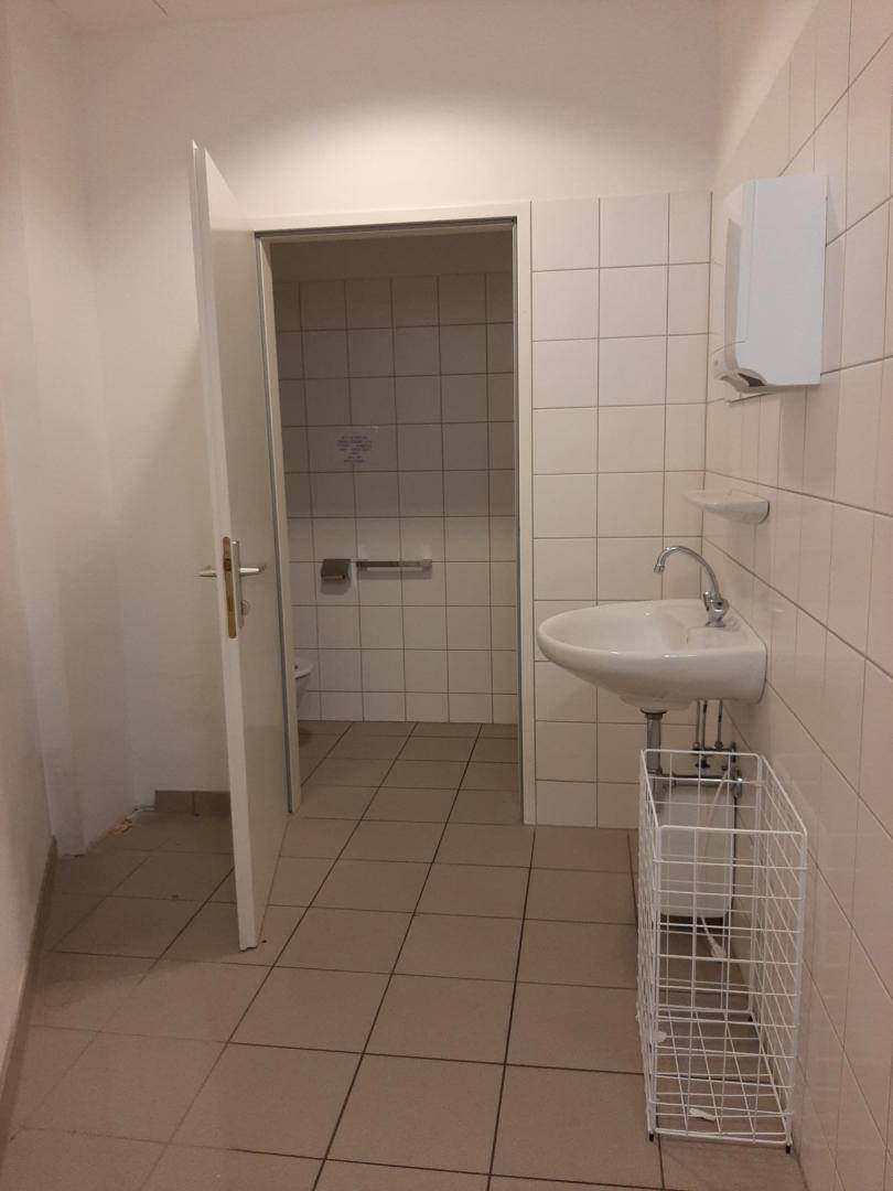 Einfaches Badezimmer mit weißen Fliesen, Waschbecken und einem offenen Türrahmen zum Toilettenbereich.
