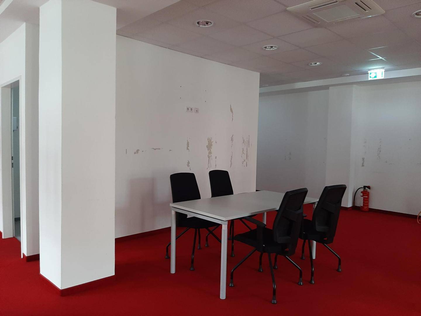 Bürobereich mit rotem Teppichboden, einem Besprechungstisch und einem Feuerlöscher an der Wand.