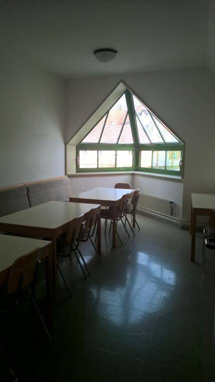 Ein Gemeinschaftsraum mit Tischen und Stühlen, beleuchtet durch ein großes dreieckiges Fenster, das eine Dachschräge andeutet.