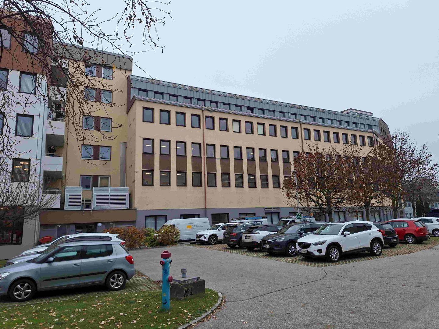 Ansicht eines mehrstöckigen Bürogebäudes mit Parkplätzen davor und Bäumen im Herbstlaub.