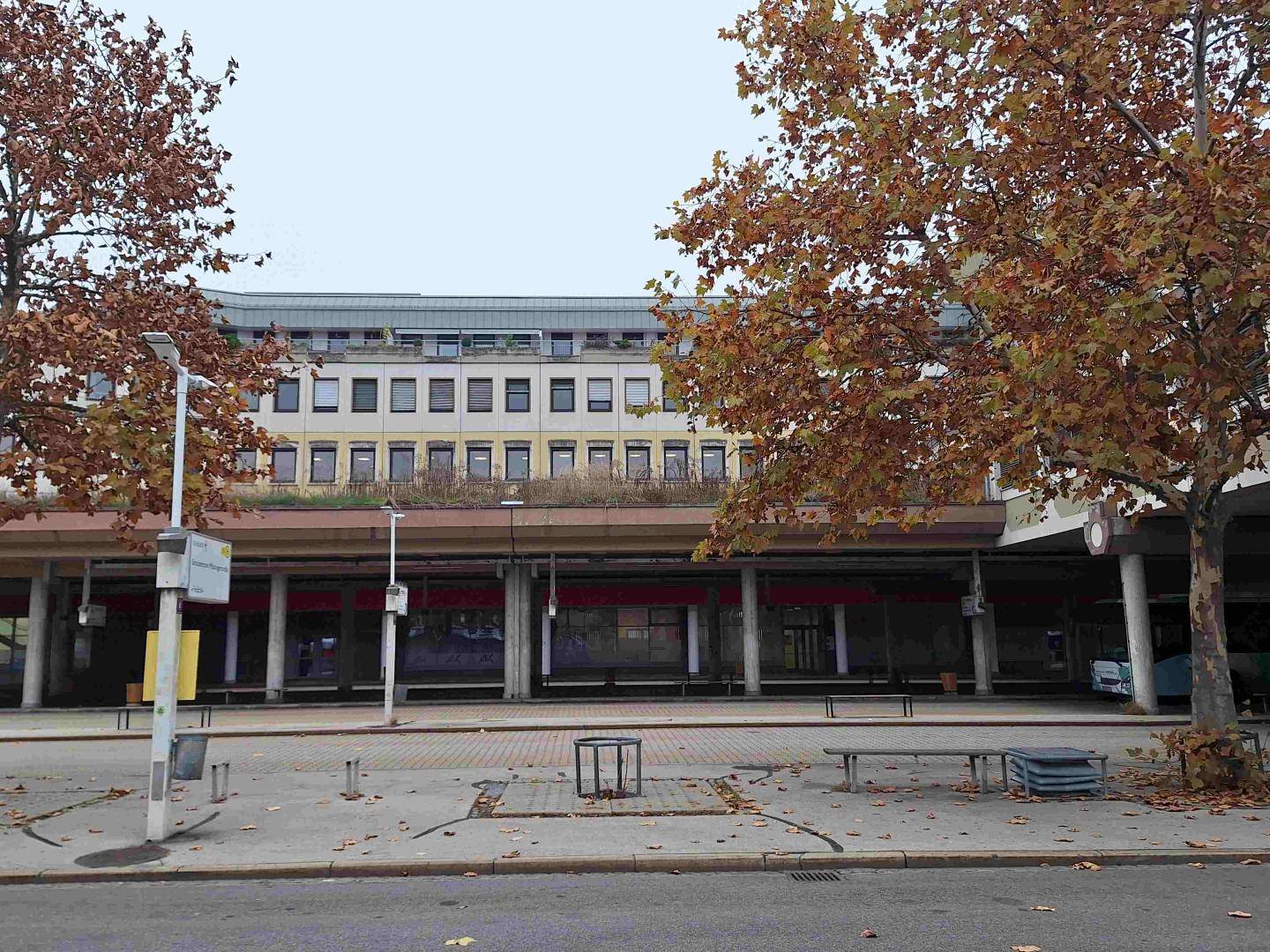 Fassade eines Gebäudes mit Bushaltestelle im Vordergrund, umrahmt von Bäumen mit Herbstlaub.