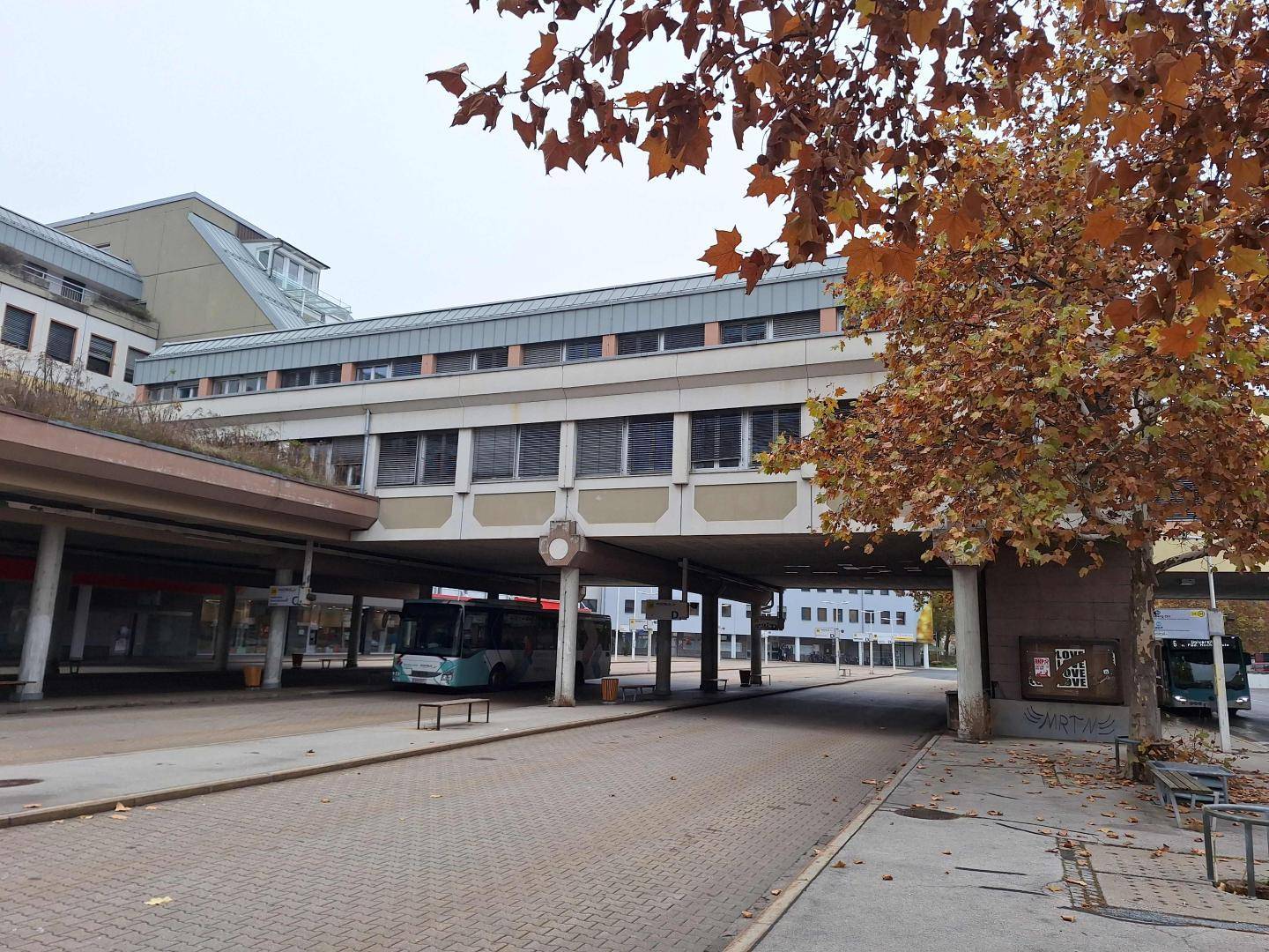 Bushaltestelle mit einem modernen Gebäude und einer Fußgängerbrücke, umgeben von Bäumen im Herbst.