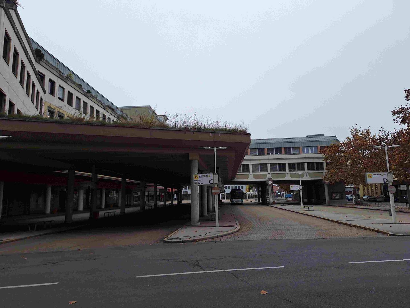 Blick auf eine Bushaltestelle mit einem modernen Gebäude und einer darüberliegenden Verbindungsbrücke.