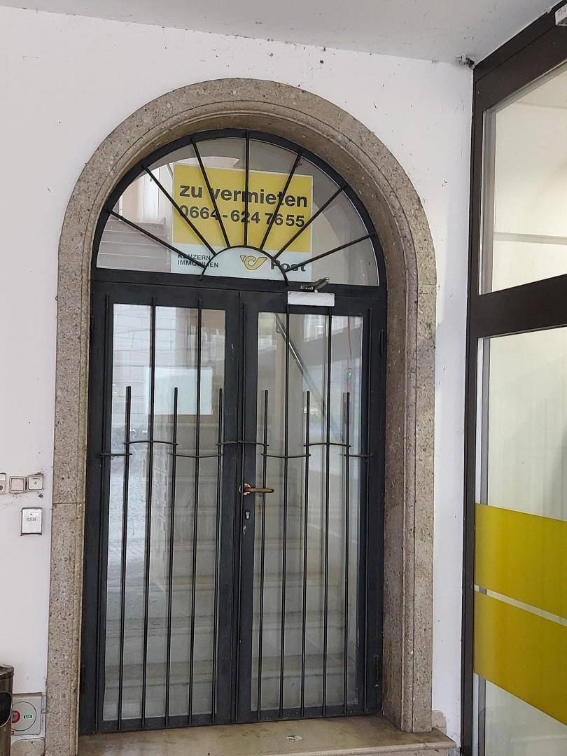 Historischer Eingang mit Rundbogen aus Stein, Glastüren und Metallgittern, mit einem 'Zu vermieten'-Schild.