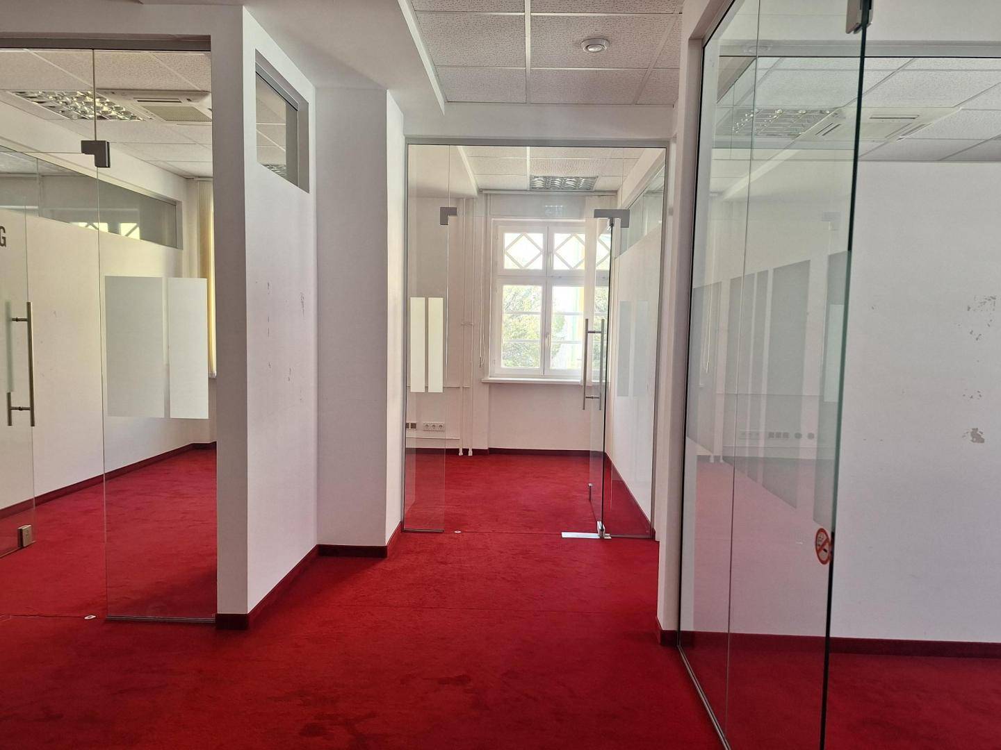Heller Bürobereich mit rotem Teppichboden und transparenten Glaswänden, die einzelne Büros abgrenzen.