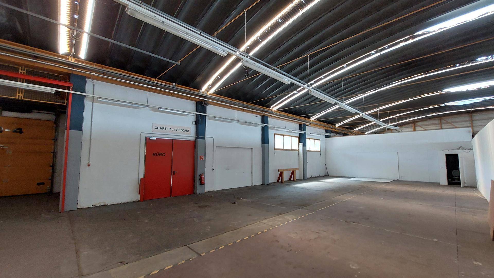 Großer, offener Bereich einer Industriehalle mit weißen Wänden, Betonboden und roten Doppeltüren.