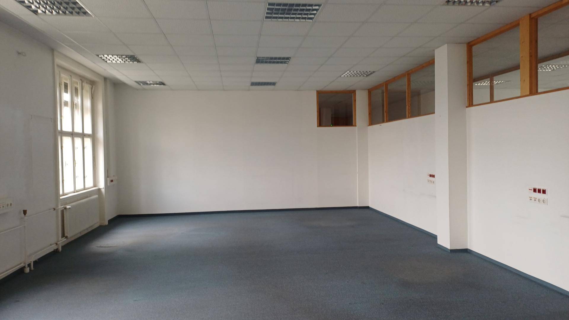 Großer, leerer Büroraum mit Teppichboden, weißen Wänden und Fenstern mit Holzrahmen.