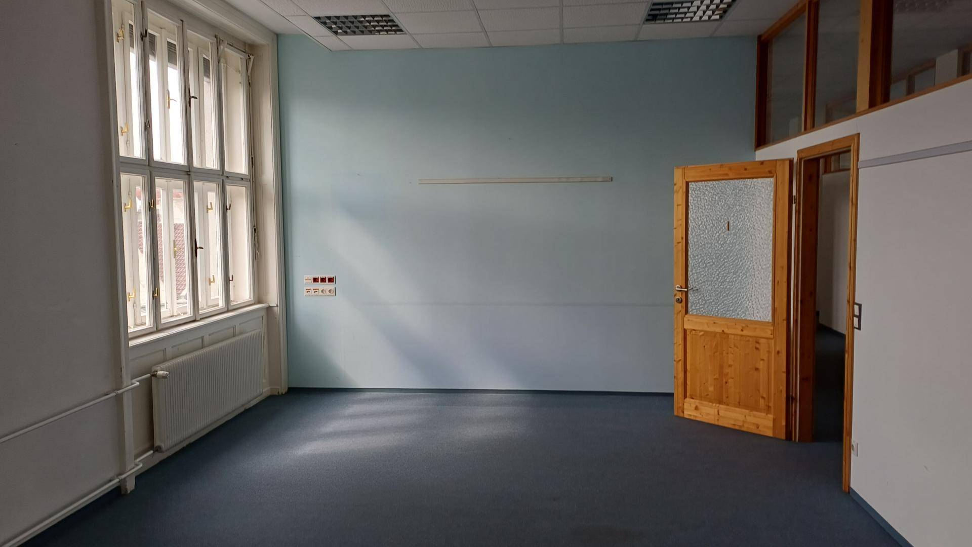 Büroraum mit hellblau gestrichener Wand, Teppichboden und einer Holztür mit Glaseinsatz.