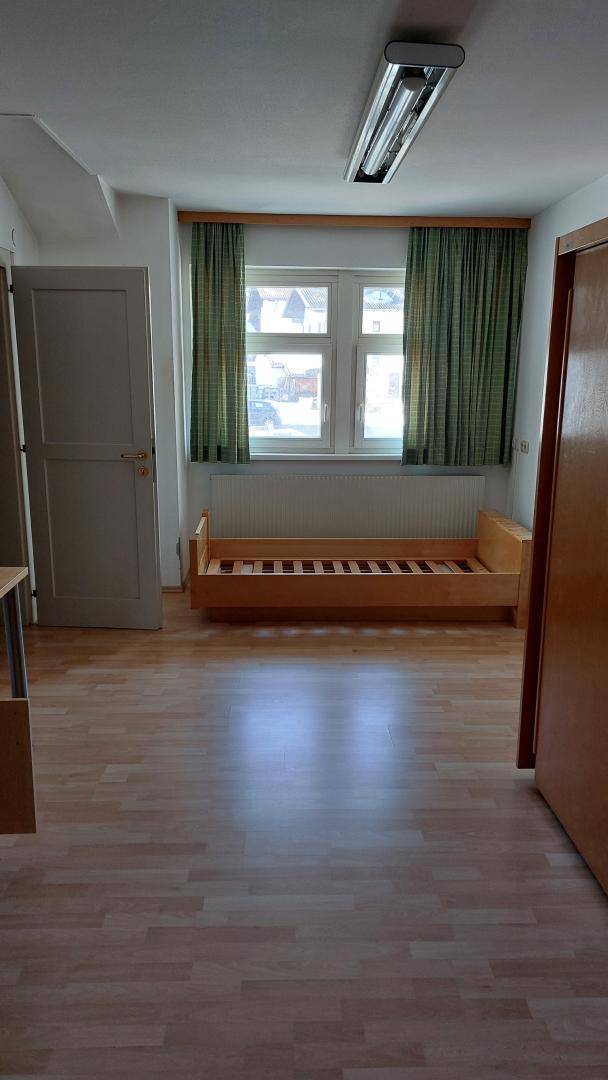 Schlafzimmer mit Laminatboden, Fenster mit Vorhängen und einem einfachen Holzbettgestell.