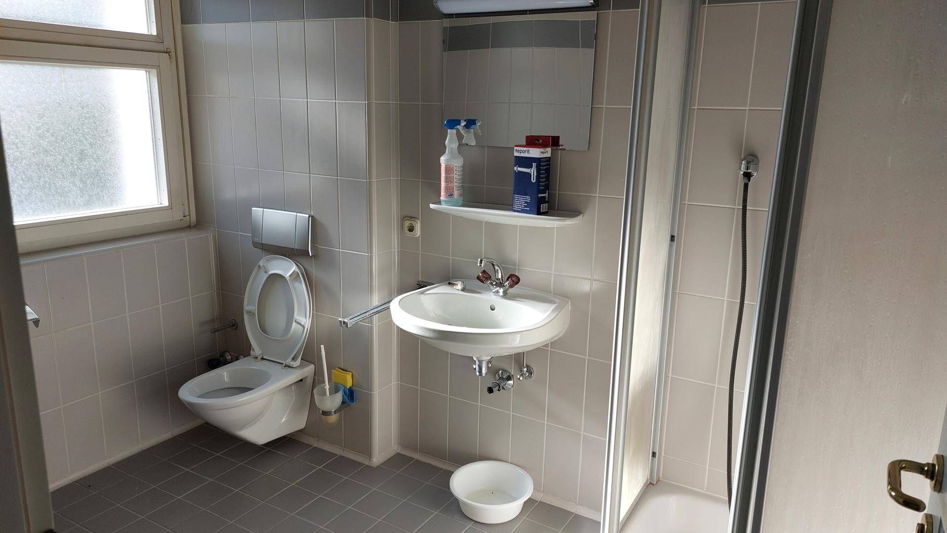 Badezimmer mit grauen Fliesen, wandhängendem WC, Waschbecken und Duschkabine.
