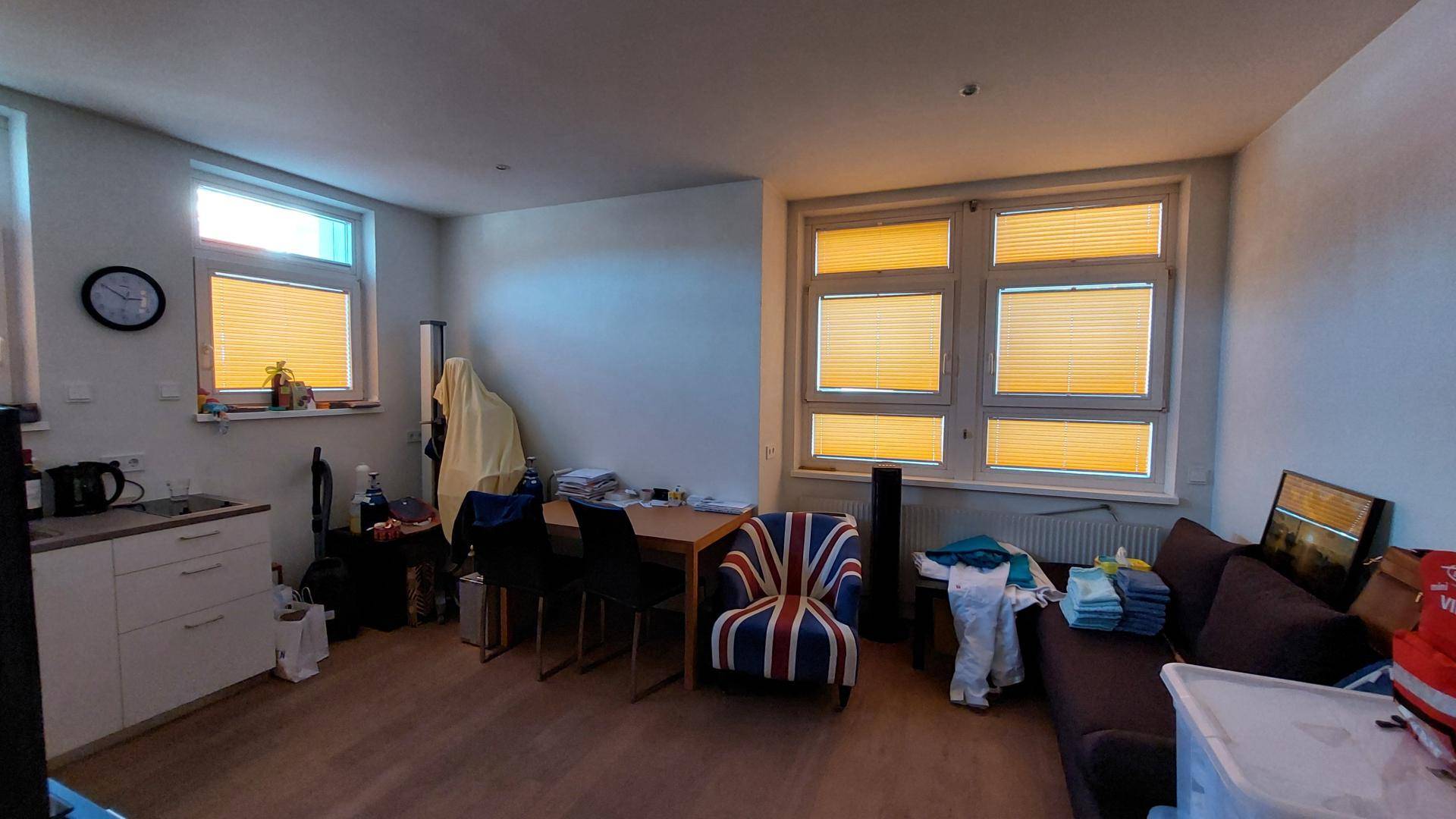 Wohn- und Essbereich mit kleiner Küchenzeile, Laminatboden und großen Fenstern mit Jalousien.