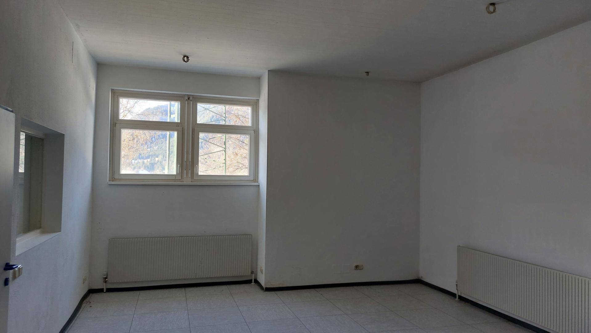 Leerer, heller Raum mit Fliesenboden, Heizkörpern und einem großen Fenster.
