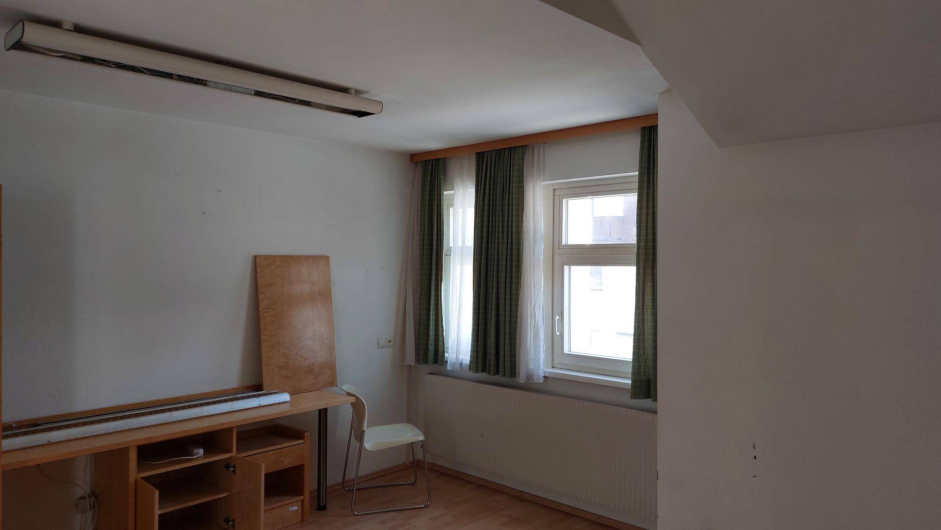 Heller Raum mit Laminatboden, Fenster mit Vorhängen und einem einfachen Schreibtisch.
