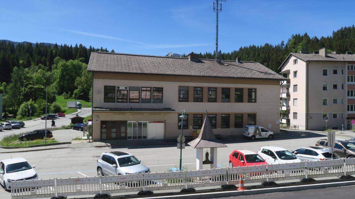 Außenansicht des Gebäudes mit großem Parkplatz, umgeben von Bäumen und Bergen unter klarem blauem Himmel.