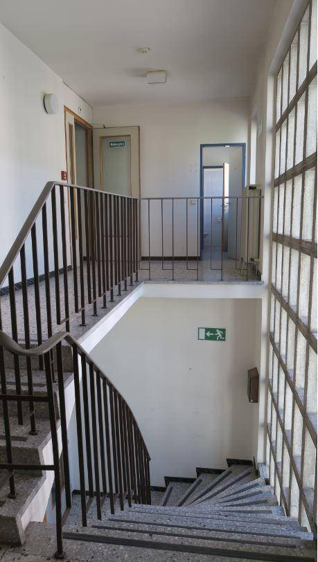 Treppenhaus mit Terrazzostufen, Metallgeländer und großen Fenstern, die viel Licht spenden.