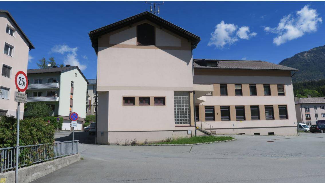Seitenansicht des Gebäudes mit heller Fassade, Glasbausteinen und umliegenden Gebäuden sowie Bergen im Hintergrund.