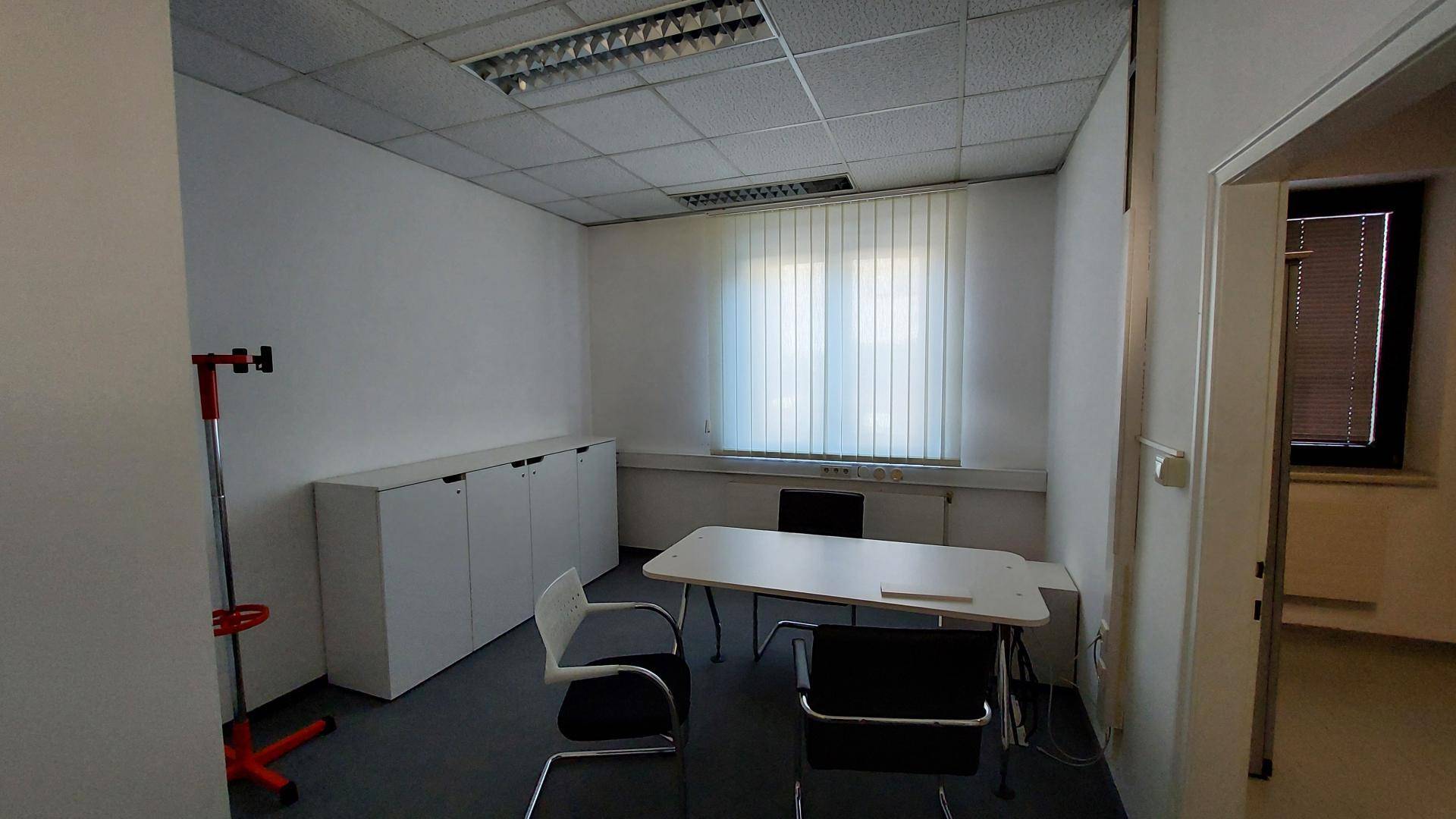 Funktional eingerichtetes Büro mit Schreibtisch, Stühlen, Schränken und einem Fenster mit Jalousien.