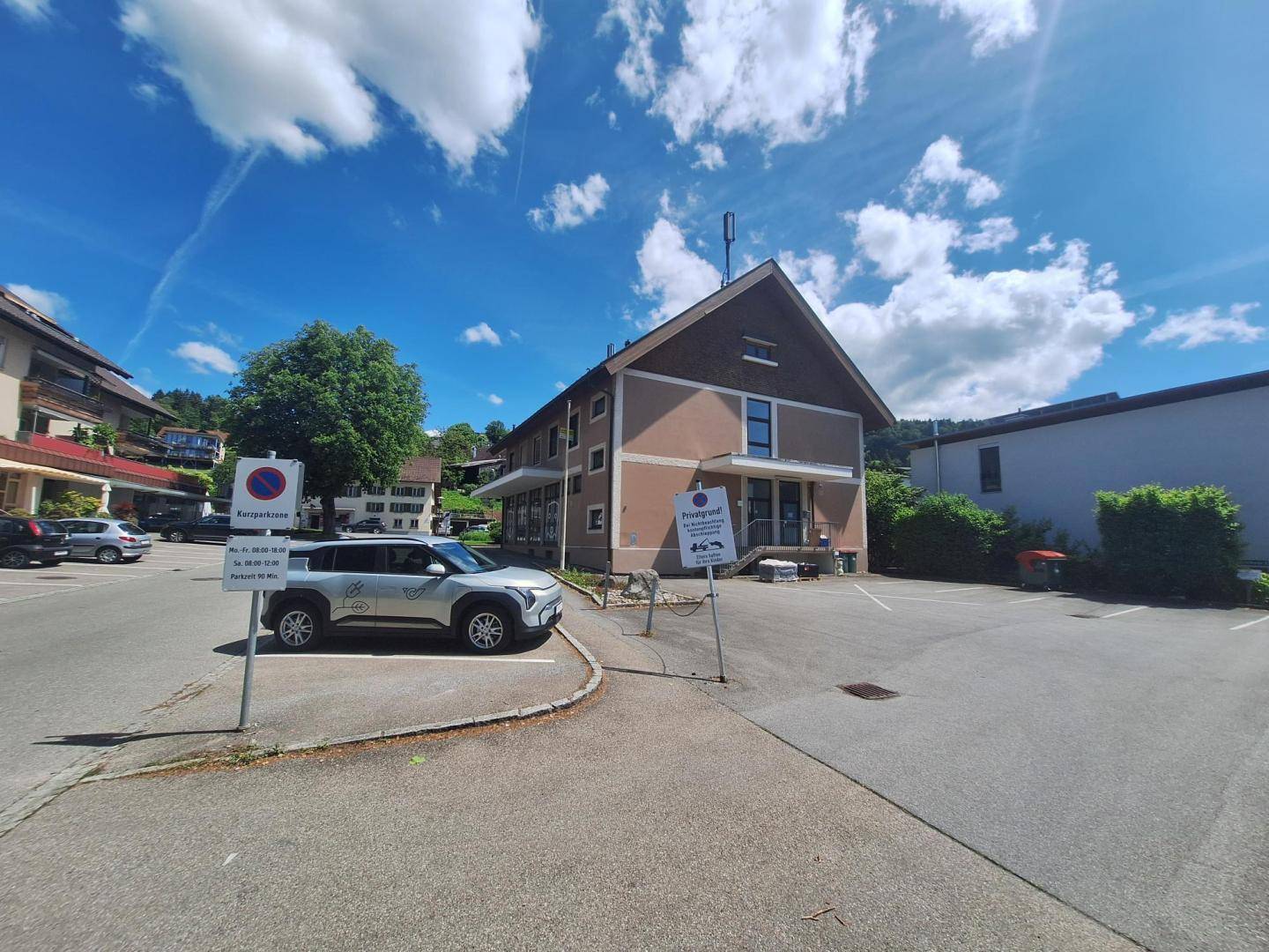 Weitwinkelansicht des Gebäudes mit Parkplatz, umliegenden Häusern und sonnigem Wetter.