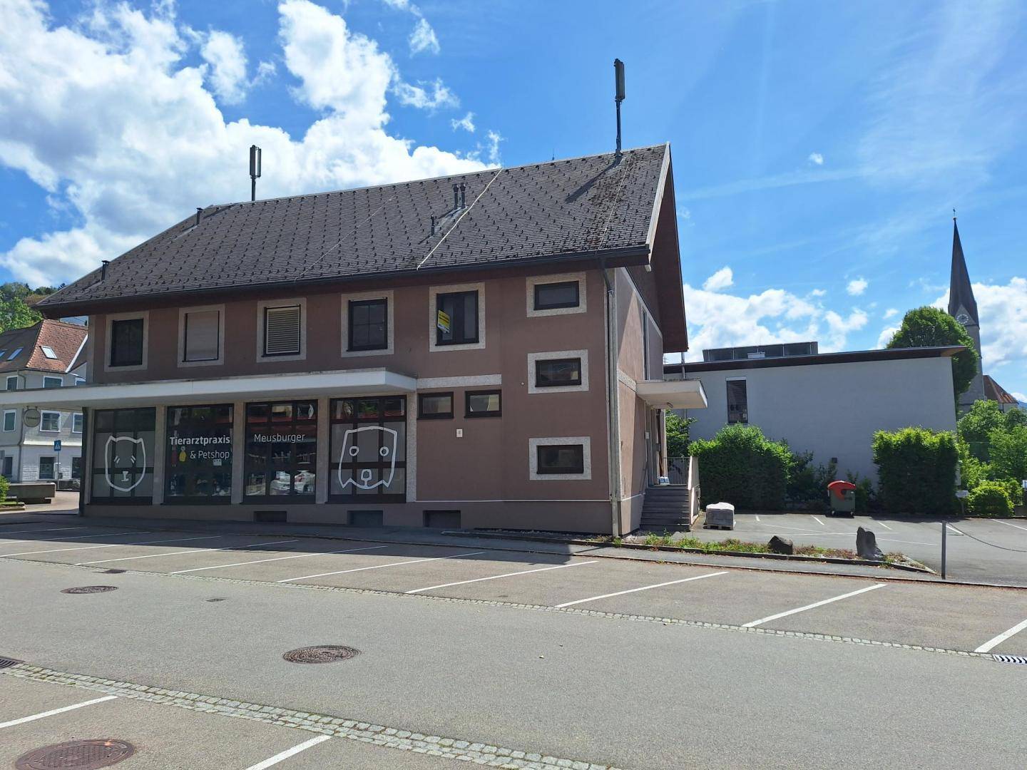 Seitenansicht des Gebäudes mit Giebeldach, Parkplatz und einem Kirchturm in der Ferne.