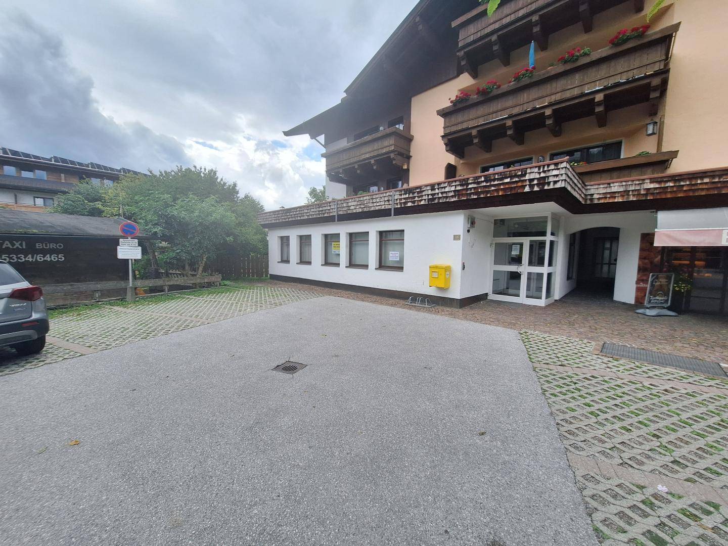 Gebäudeaußenansicht mit Parkplatz und Eingangsbereich, umgeben von Bäumen und Sträuchern.