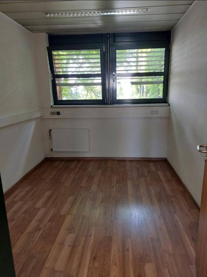 Kleiner, leerer Raum mit Laminatboden, weißen Wänden und einem Fenster mit Jalousien.