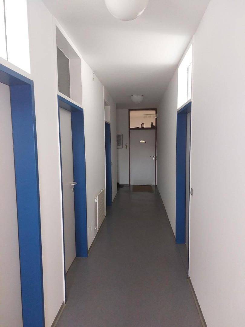 Langer, heller Flur mit weiß gestrichenen Wänden, blauen Türrahmen und grauem Vinylboden.