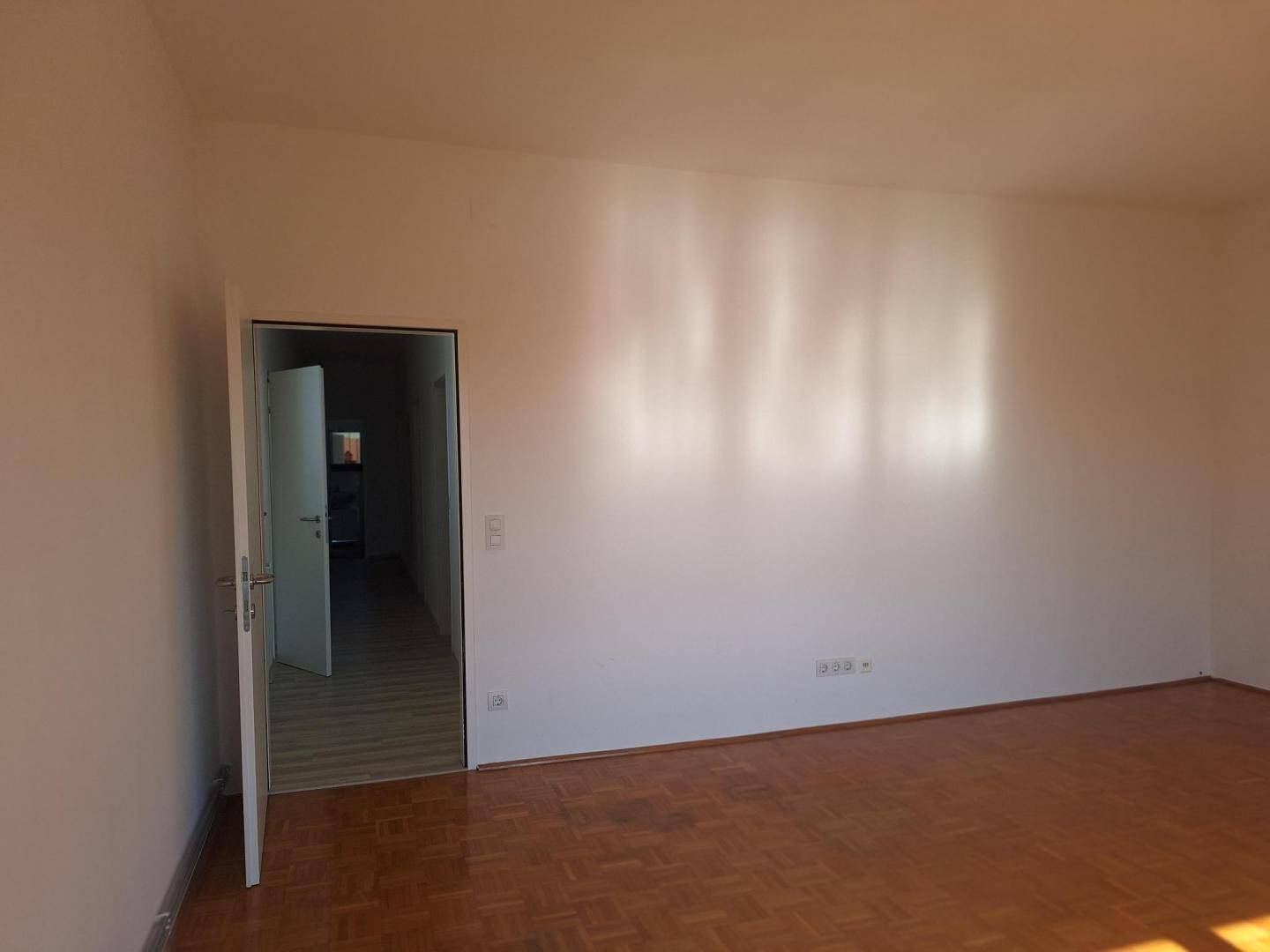 Geräumiges Wohnzimmer mit Parkettboden und Zugang zu einem weiteren Raum.