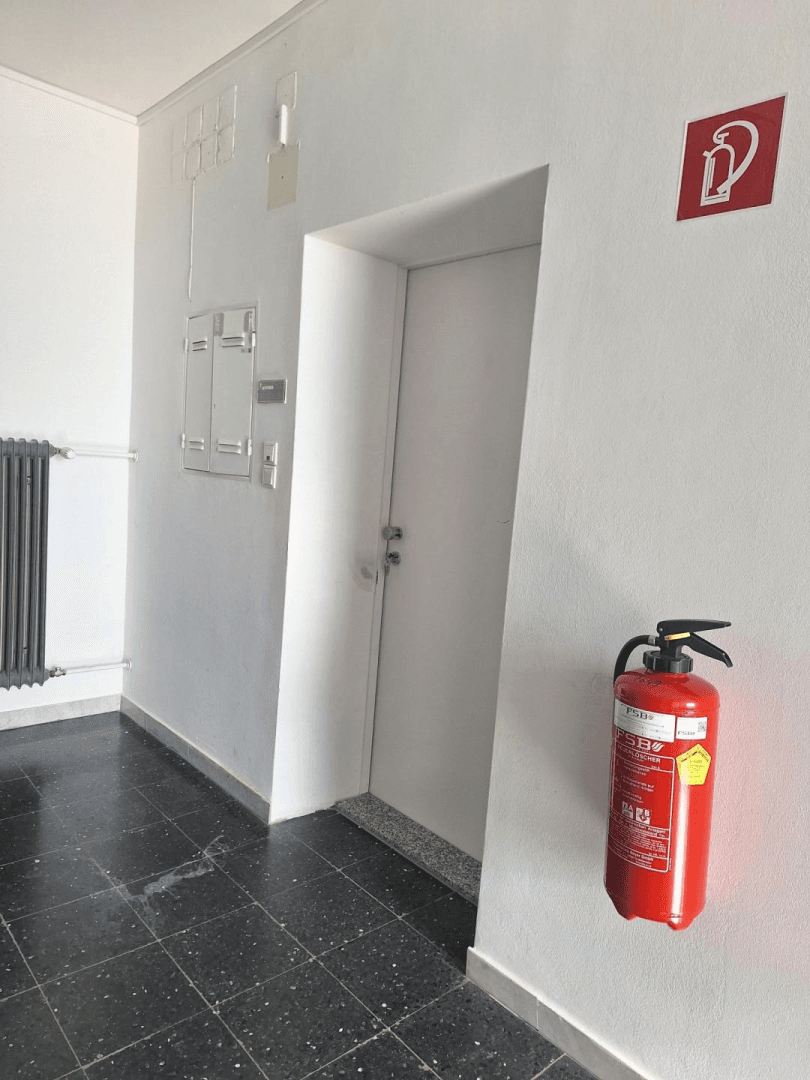 Heller Flur mit weiß gestrichenen Wänden, dunklem Boden und Feuerlöscher.