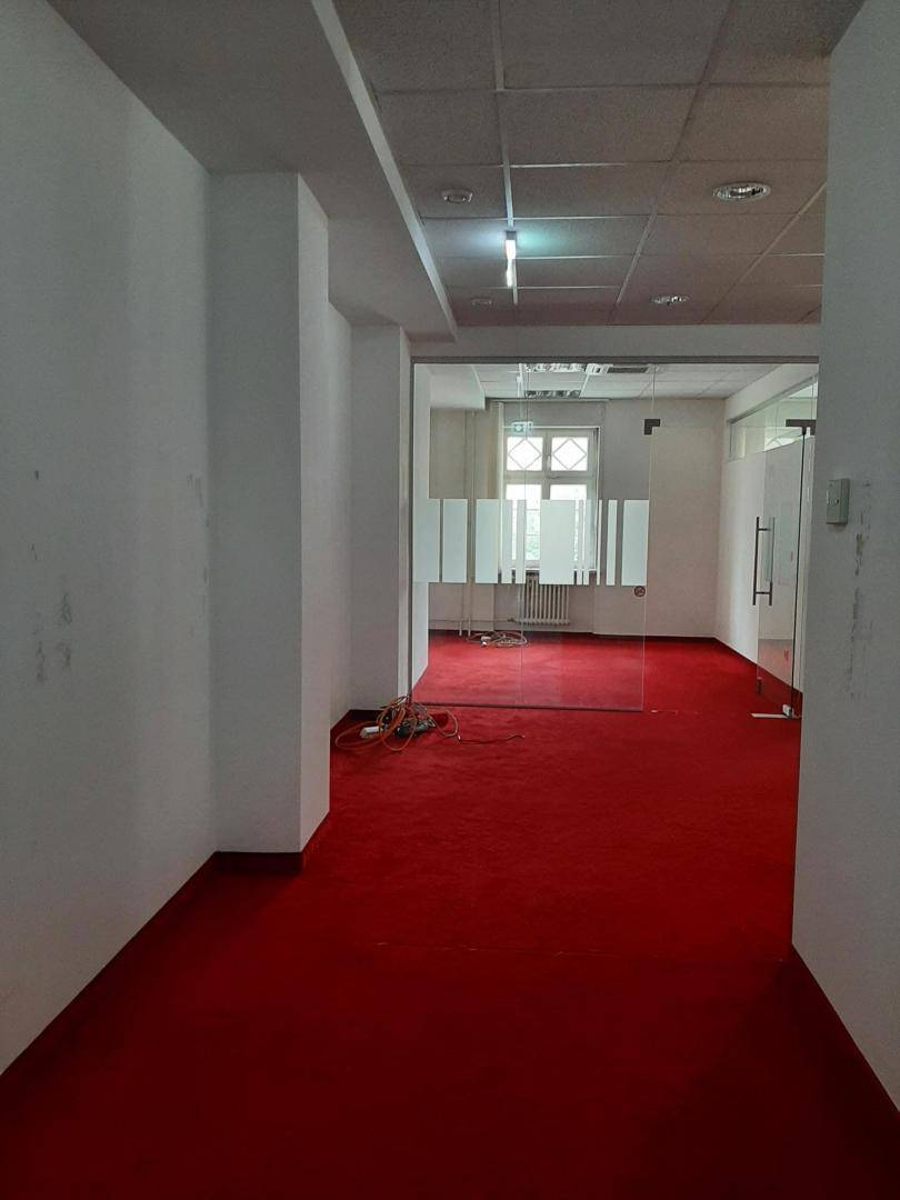Langer Bürogang mit rotem Teppichboden und Blick in einen Raum mit Glaswänden und Fenstern.