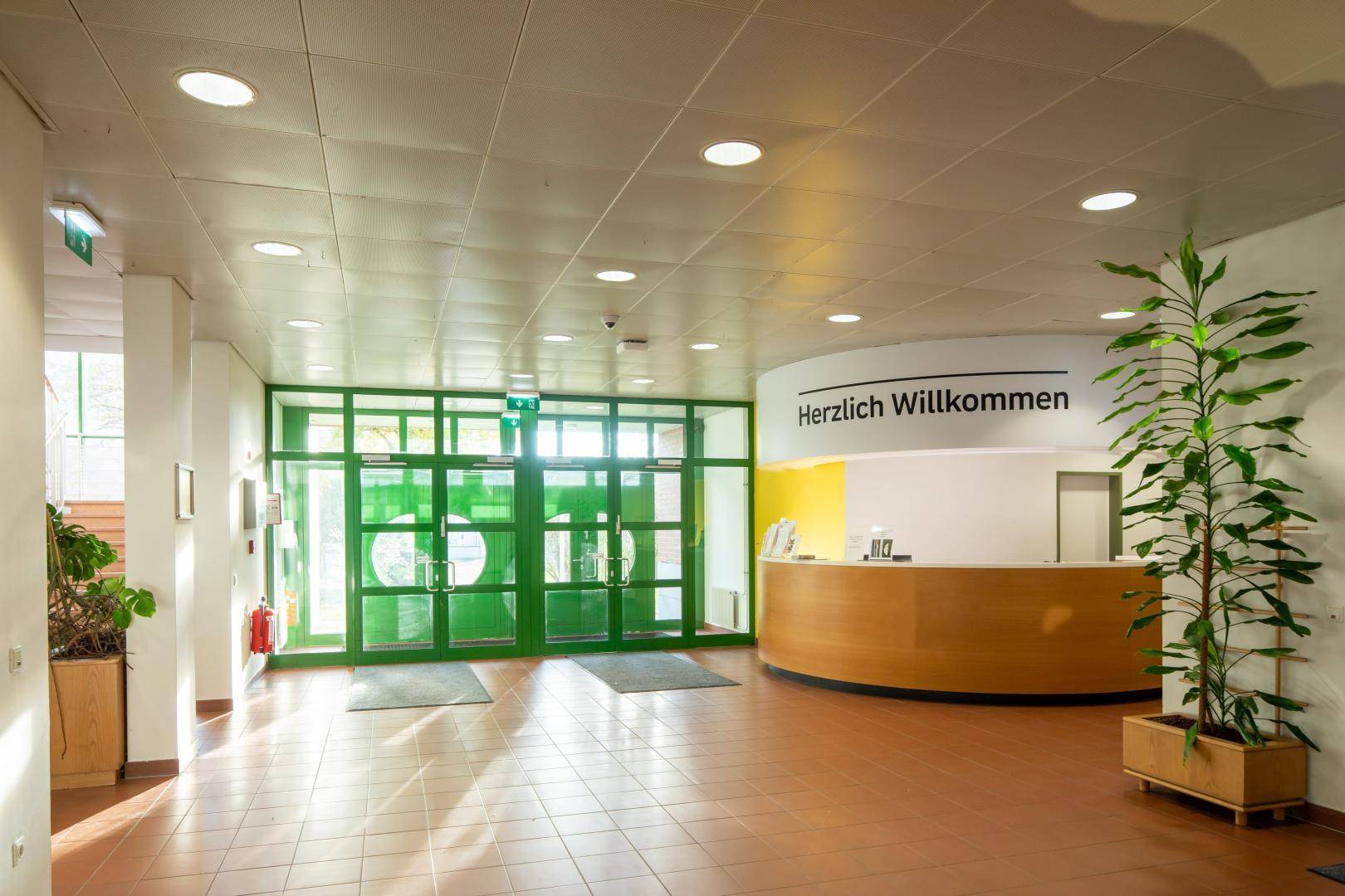 Heller Empfangsbereich mit Willkommensschild, Holztheke und großen grünen Glastüren zum Eingang.