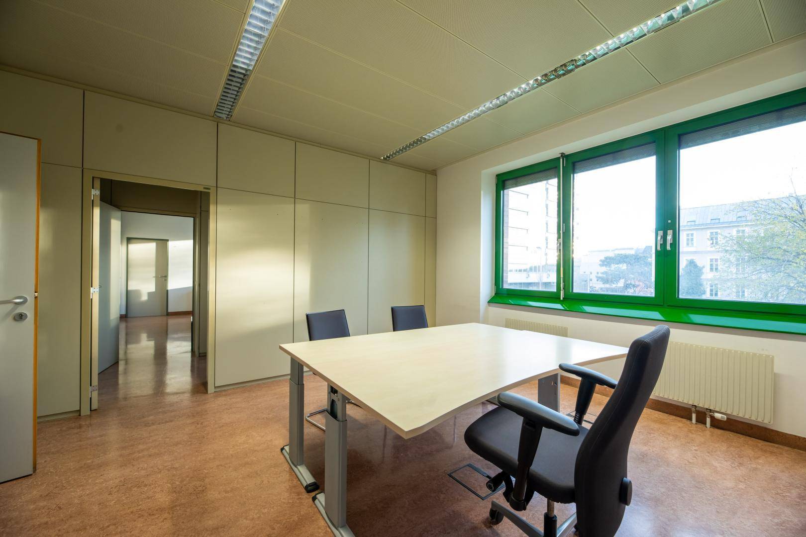 Helles Büro mit großem Fenster, Besprechungstisch und Zugang zu einem weiteren Raum.