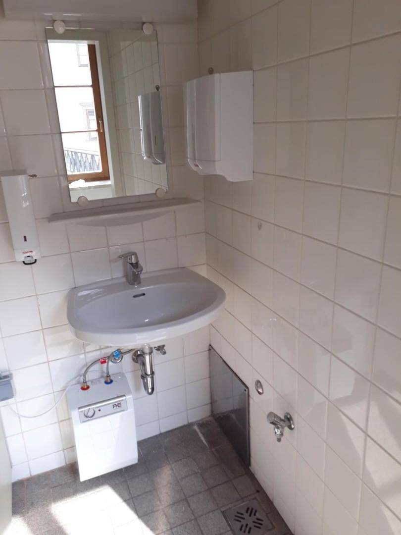 Kleines Badezimmer mit weißen Fliesen, Waschbecken und einem Fenster für natürliches Licht.