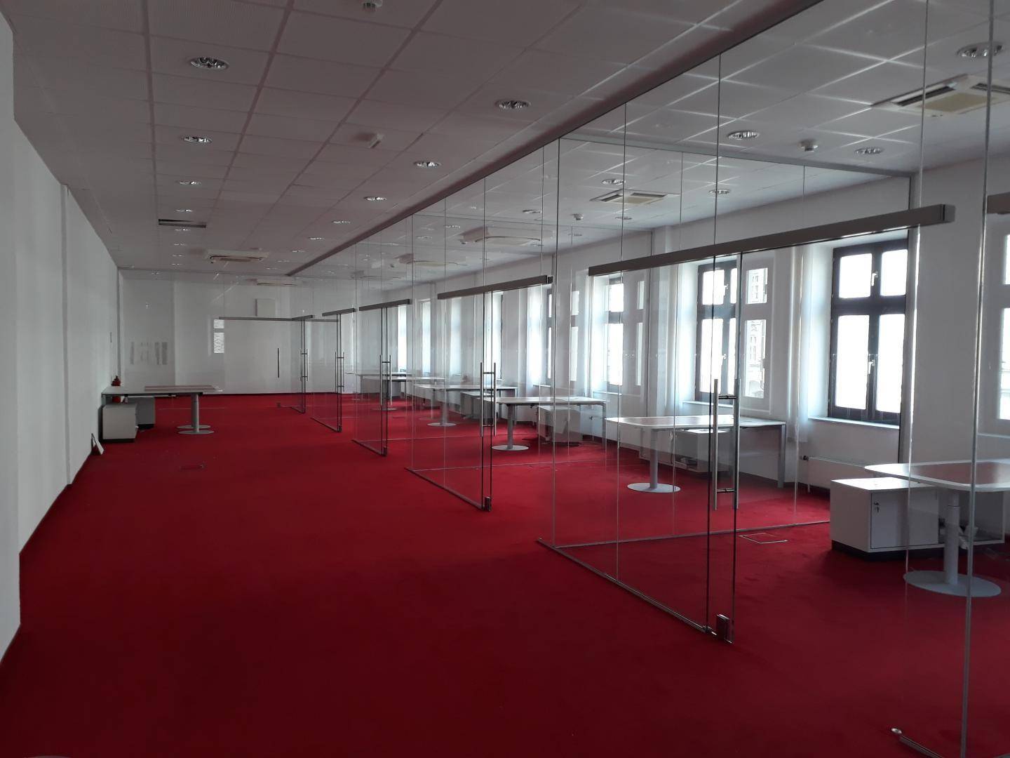 Großes, leeres Büro mit rotem Teppichboden, Glasabtrennungen und vielen Fenstern für Tageslicht.