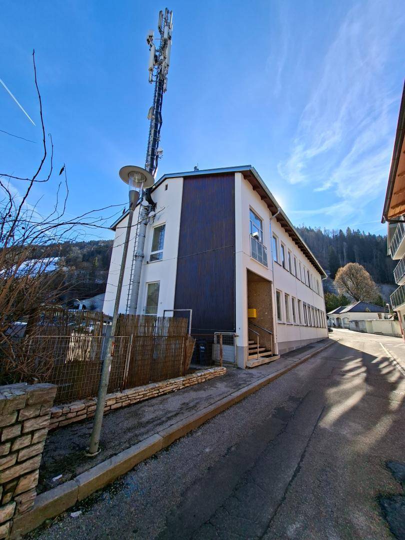 Gebäudeansicht von der Seite mit dunkler Verkleidung und weißer Fassade, Mobilfunkmast und Straße.