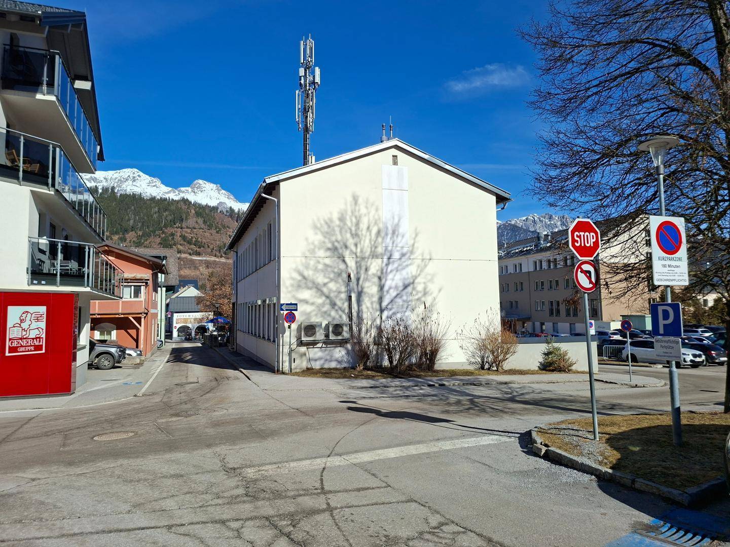 Vorderansicht des Gebäudes mit weißer Fassade, Mobilfunkmast und klarer Bergsicht im Hintergrund.