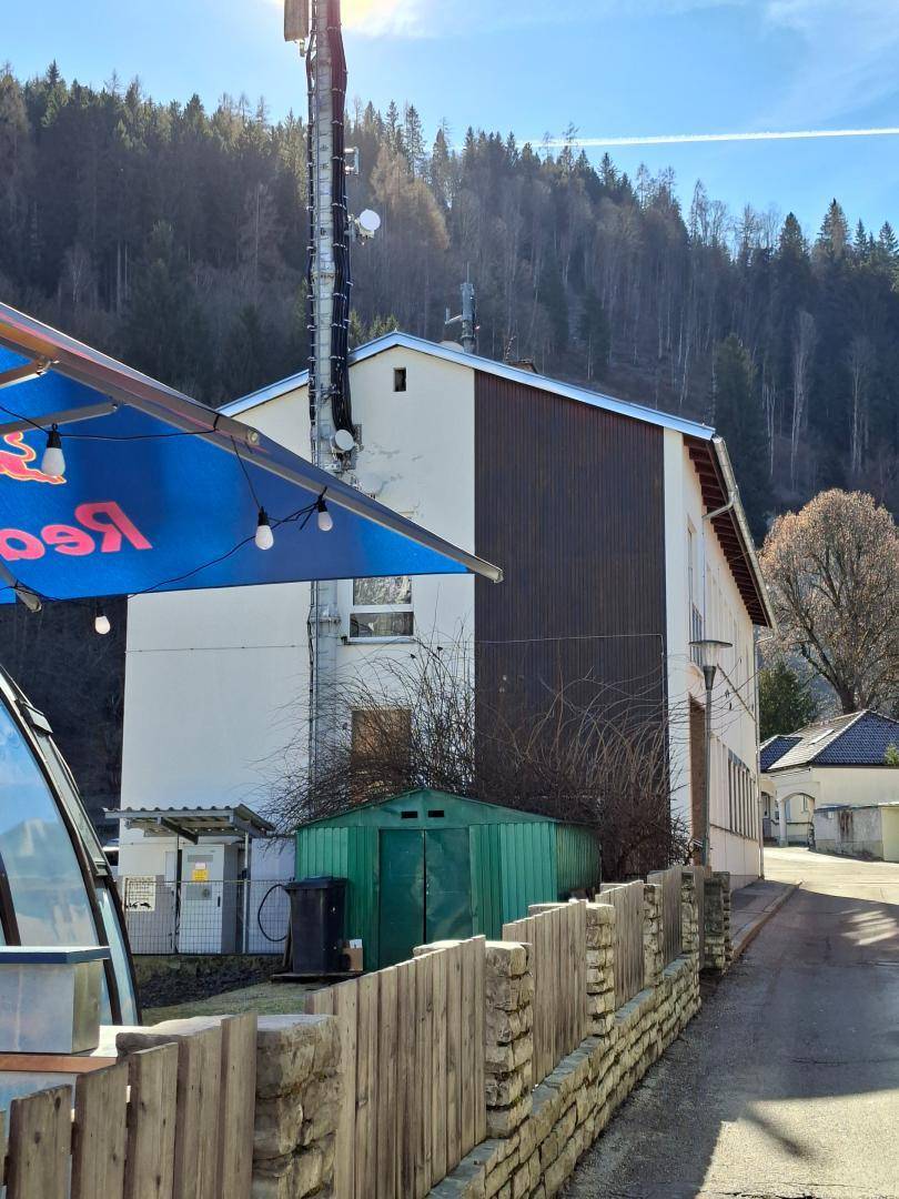 Rückansicht des Gebäudes mit grünem Schuppen, Holzzaun und Mobilfunkmast vor bewaldetem Hang.