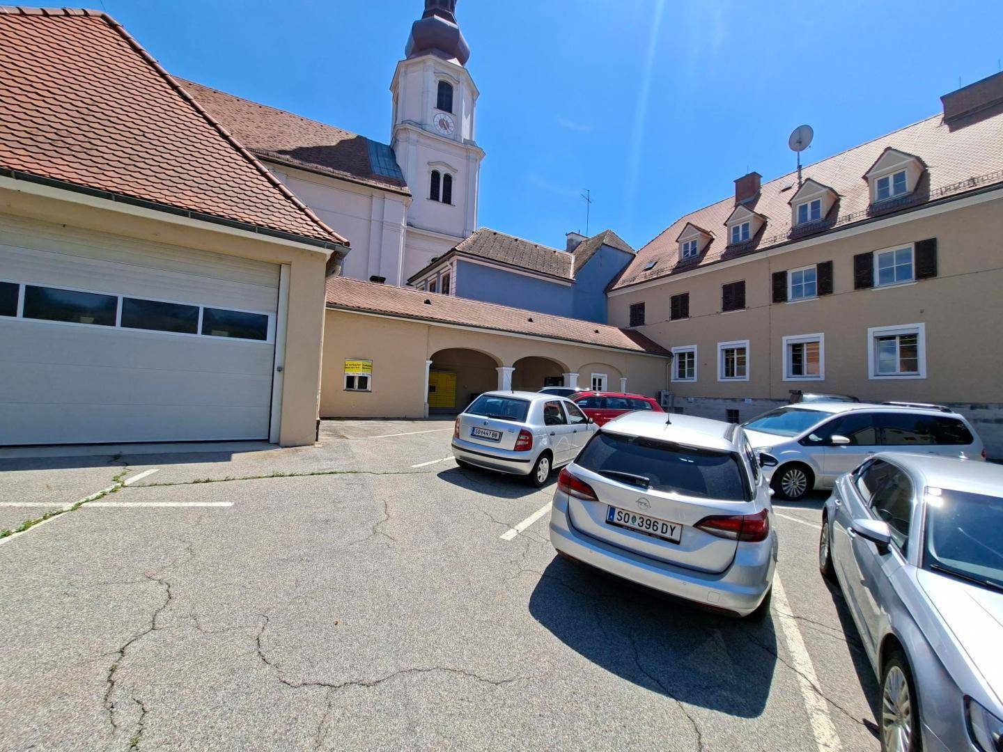 Blick auf den Parkplatz mit geparkten Fahrzeugen, umgeben von historischen Gebäuden und einem Kirchturm.