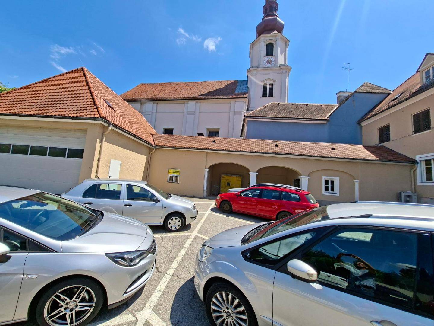 Parkplatz mit mehreren Autos vor einer historischen Kirche und angrenzenden Gebäuden mit Garagentor.