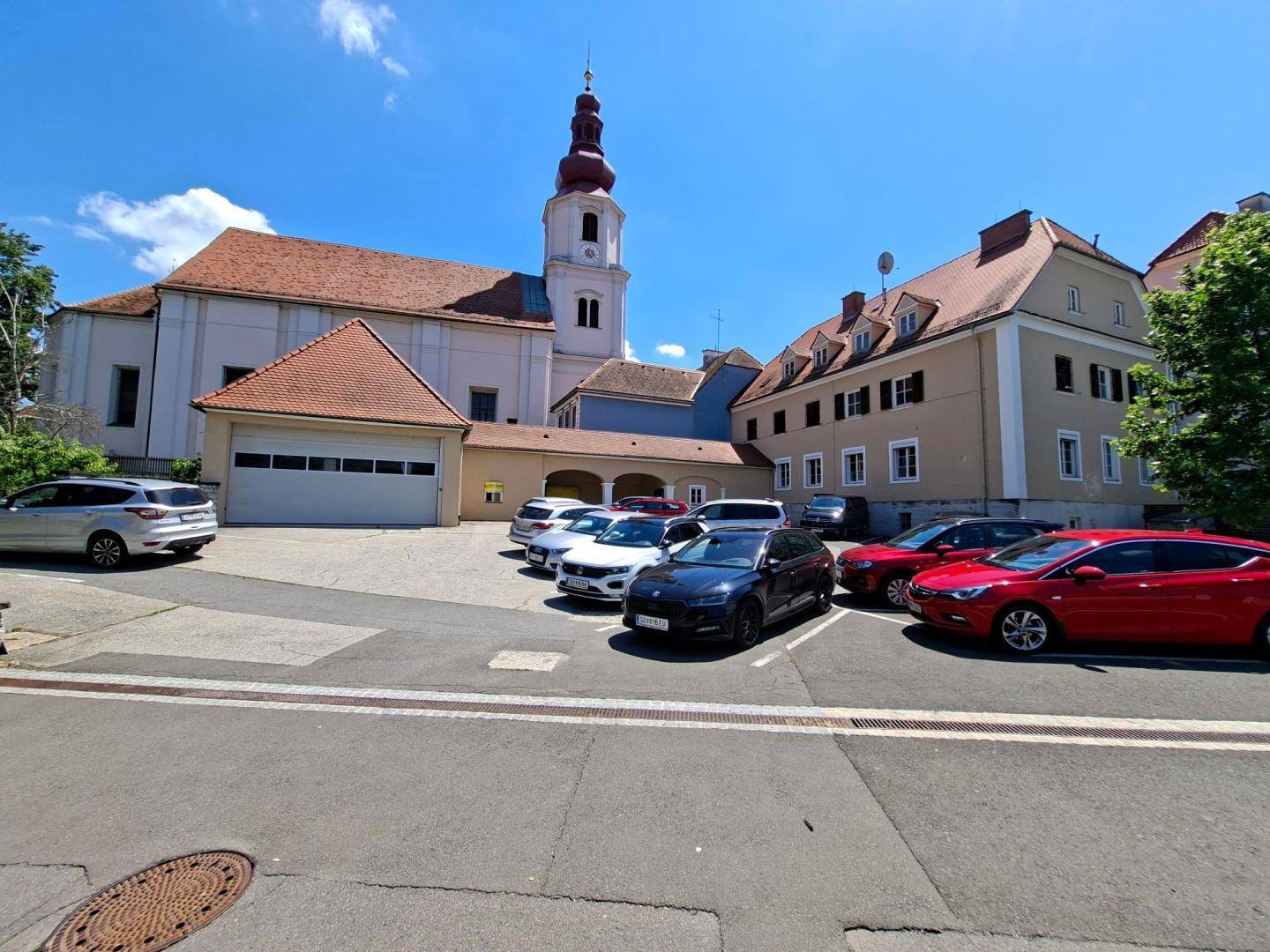 Weitläufiger Blick auf einen Parkplatz vor historischen Gebäuden und einer Kirche unter blauem Himmel.