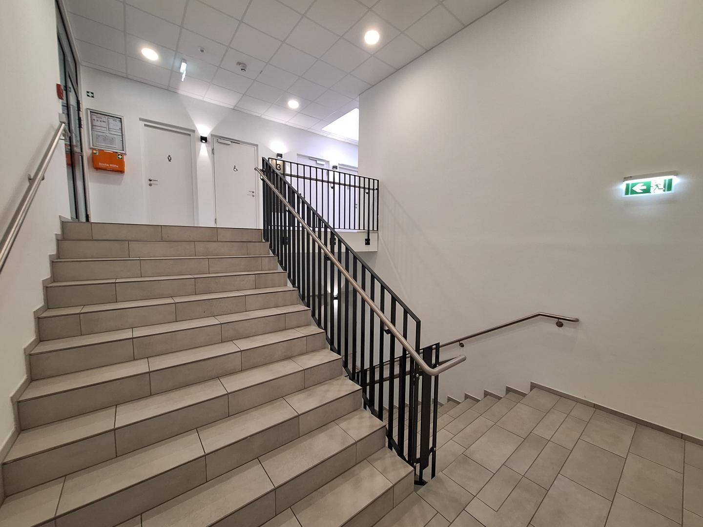 Modernes Treppenhaus mit Fliesenboden, Metallgeländer und heller Beleuchtung, führt zu oberen Etagen.