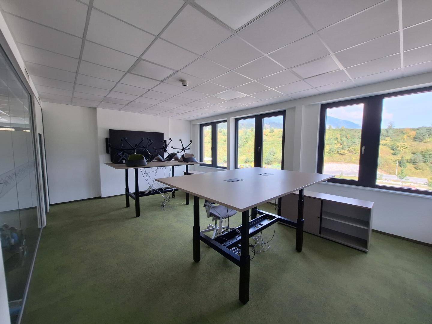 Geräumiges Büro mit grünen Teppichboden, höhenverstellbaren Tischen und großen Fenstern mit Landschaftsblick.