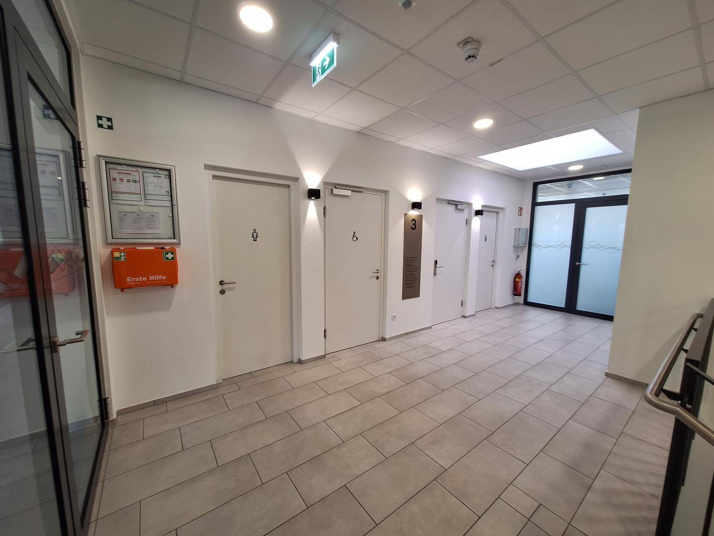 Heller Flur mit Fliesenboden, mehreren Türen und Notfallausrüstung an der Wand.