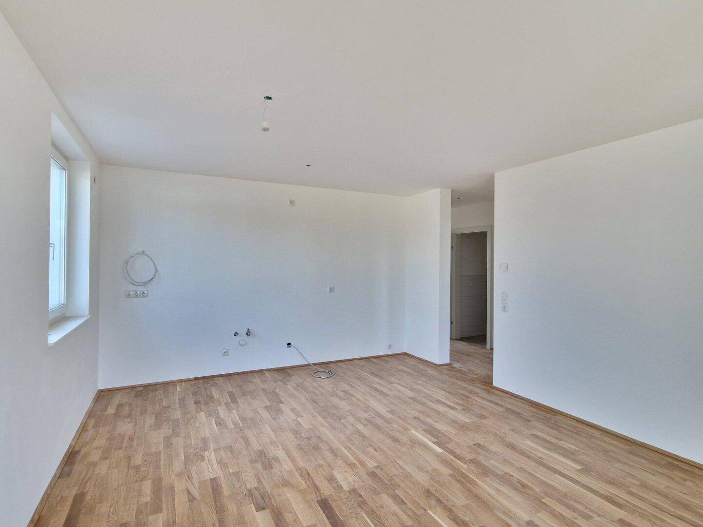 Heller, unmöblierter Wohnraum mit Parkettboden und vorbereiteten Anschlüssen für eine Küche.