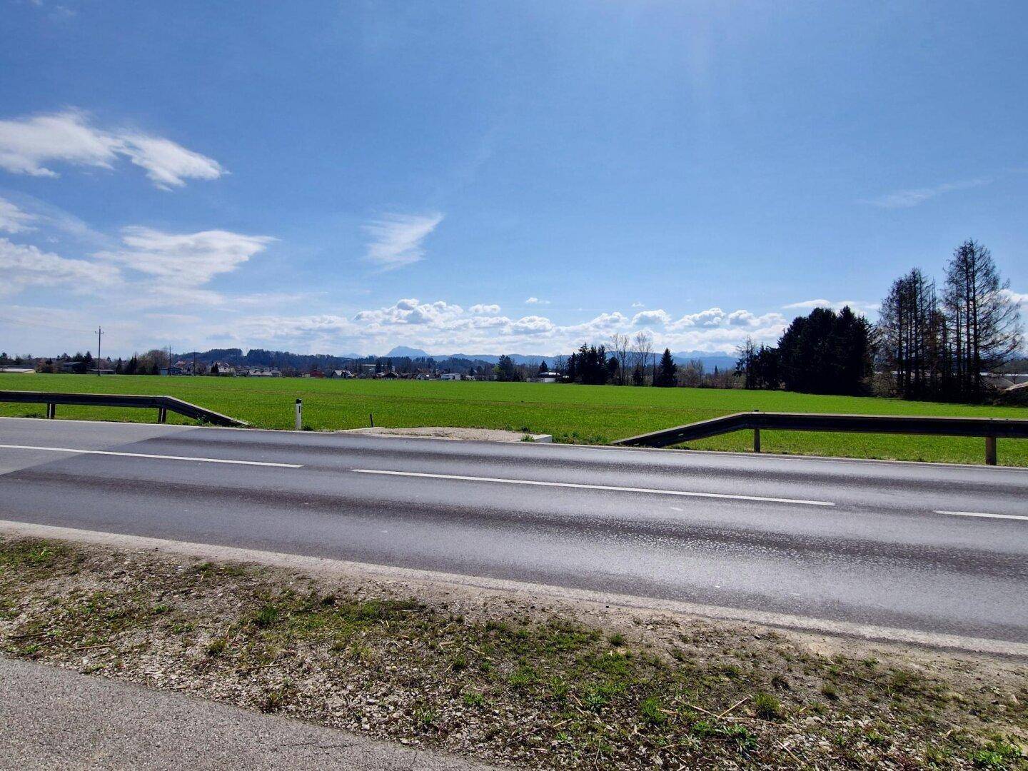 Landstraße neben einer grünen Wiese mit Bergen und Häusern in der Ferne unter blauem Himmel.