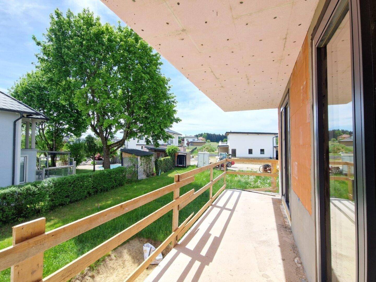 Balkon im Rohbau mit Holzgeländer und Blick auf die grüne Umgebung und Nachbargebäude.